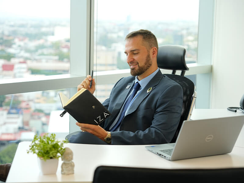 Hombre sonriente trabajando en laptop junto a ventanal con planta – Iza Office Club