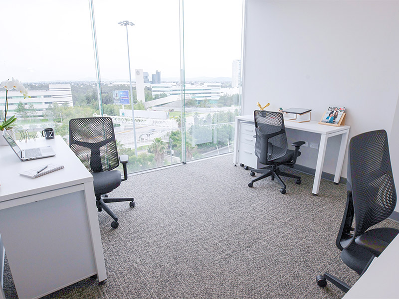 Espacio de oficina compartido con múltiples escritorios blancos, sillas y grandes ventanales con vista urbana en Inxignia, oficina en renta en Puebla