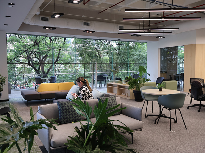 Área de lounge con sofás, plantas y mesas junto a ventanal – Iza Office Club