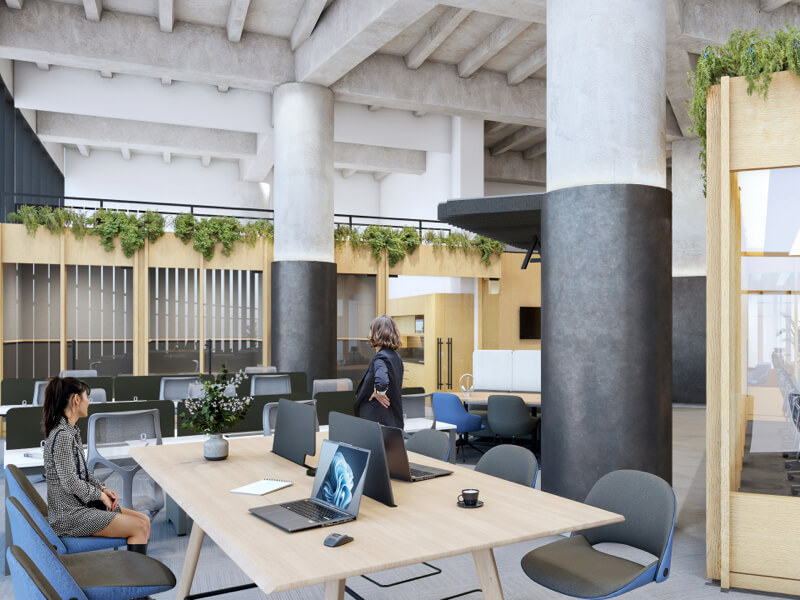 Fotografía del interior de IZA Urban Center Cancún, mostrando una amplia área de coworking con mesas de trabajo de madera, laptops y cubículos, con la presencia de dos mujeres, una sentada y otra de pie, rodeadas de grandes columnas de concreto.