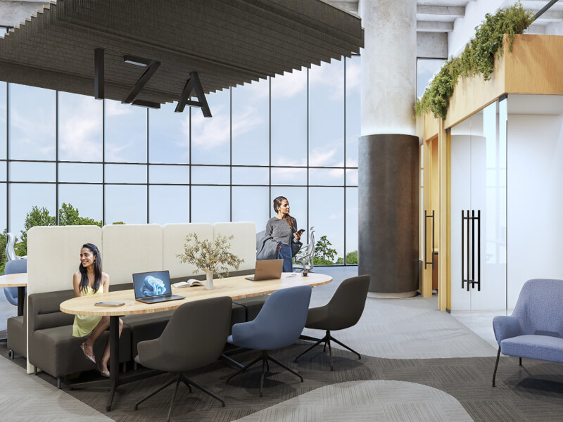 Fotografía del interior de IZA Business Centers Cancún, mostrando un área de coworking moderna con dos mujeres, una sentada sonriendo y otra de pie al fondo. El espacio está equipado con una mesa de trabajo ovalada, laptops, sillas ergonómicas y un sillón, complementado con grandes ventanales y las letras de 