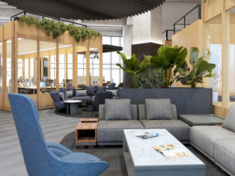Fotografía del interior de IZA Urban Center Cancún, mostrando un área común de coworking con cómodos sillones grises, un sillón individual azul y mesas de centro, decorada con plantas tropicales y cubículos de oficina con estructuras de madera al fondo.